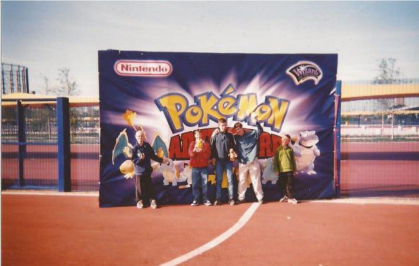 A photo of Paul and his friends standing in front of a sign with various Pokémon on it
