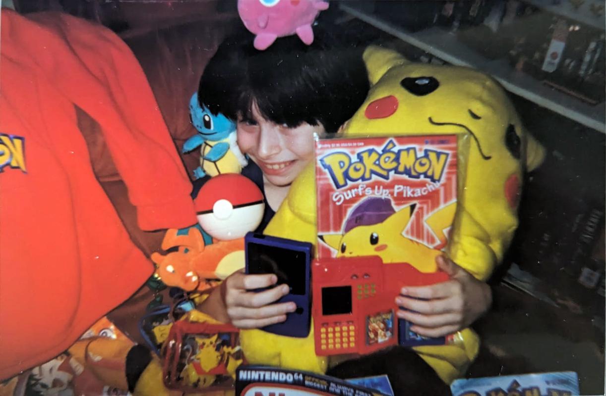 A photograph of Dazz when he was younger, holding copies of Pokémon Red & Blue, a Game Boy Color, a Pokédex toy, and a Pokémon comic called Surf's Up, Pikachu. He's surrounded by several other Pokémon related items!